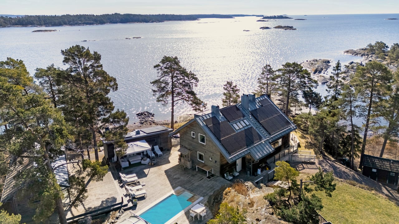 Hus med pool och trädäck bland tallar vid solglittrande hav, sett från luften.