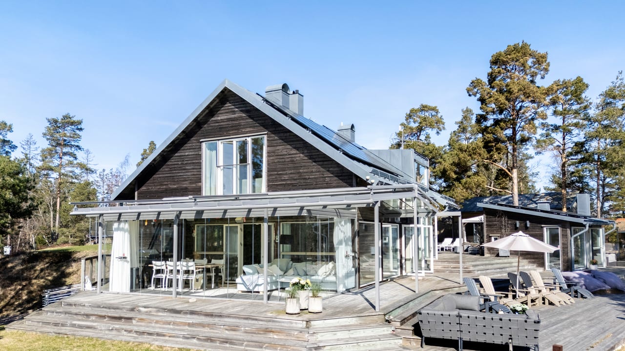 Mörkt trähus med glasad veranda och brett trädäck i sol.