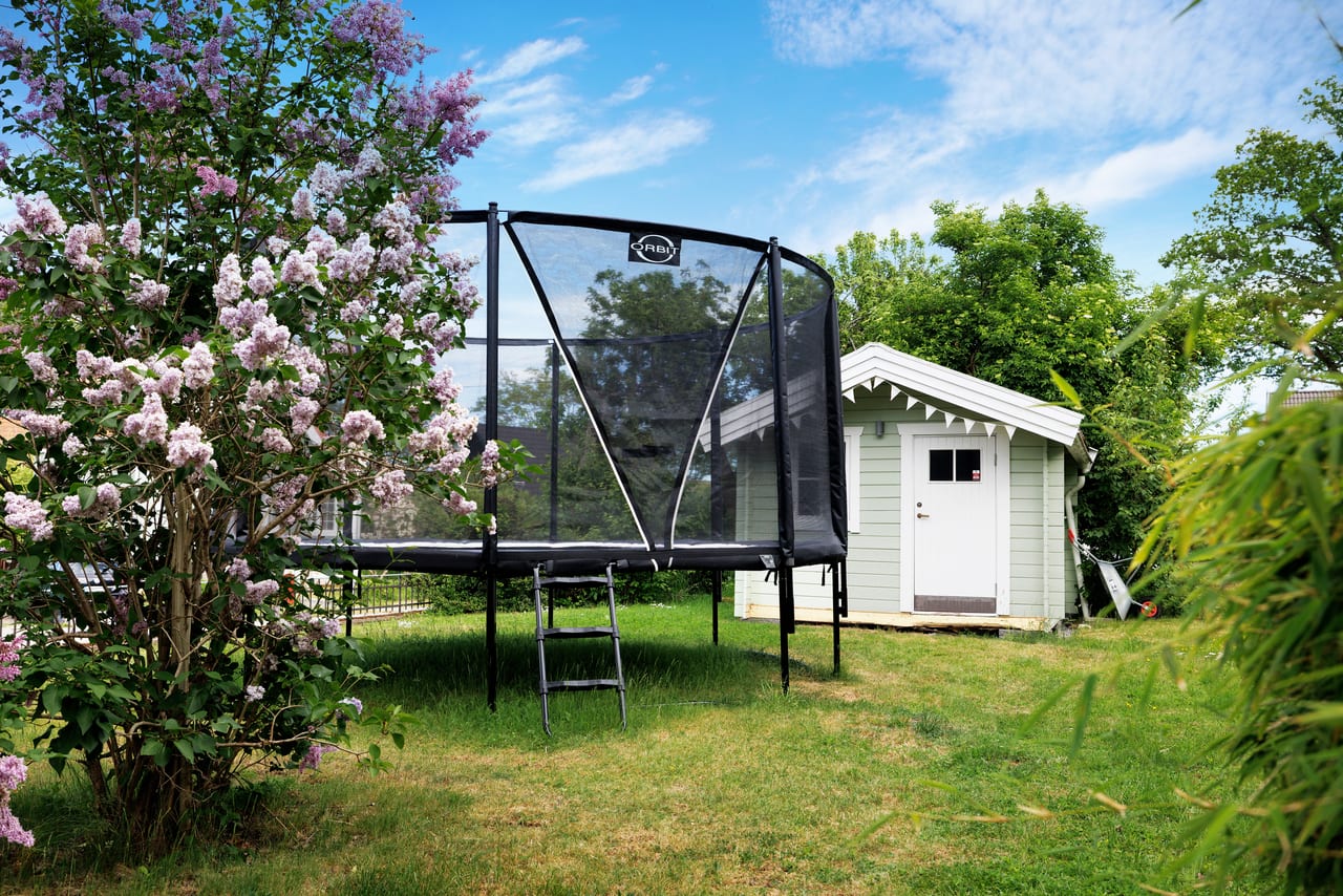 Trampolin och lekstuga på en gräsmatta framför en blommande syrenbuske.