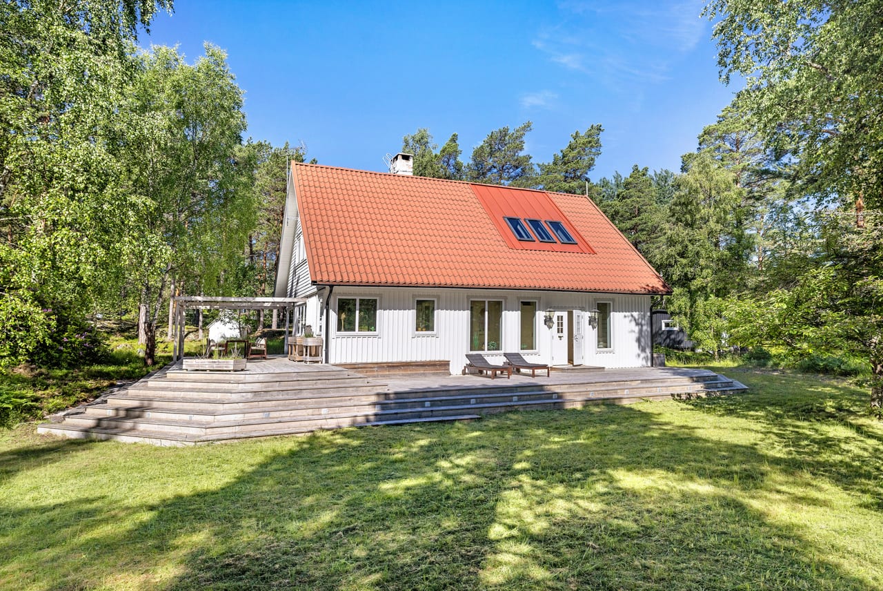 Vitt trähus med rött tegeltak och trädäck framför en gräsmatta.